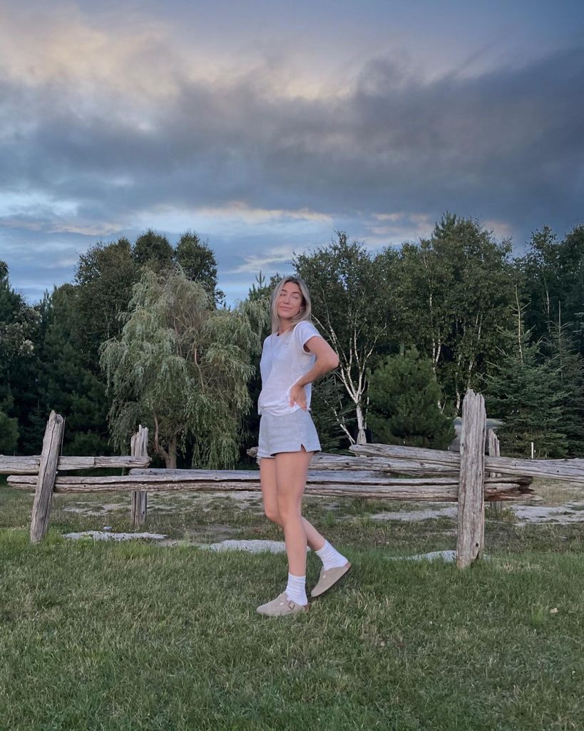 Kamilla wearing light grey shorts and white shirt posing on a field. 